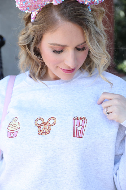 Person wearing a white sweatshirt with embroidered designs, including a popcorn bucket, scissors, and a cupcake.