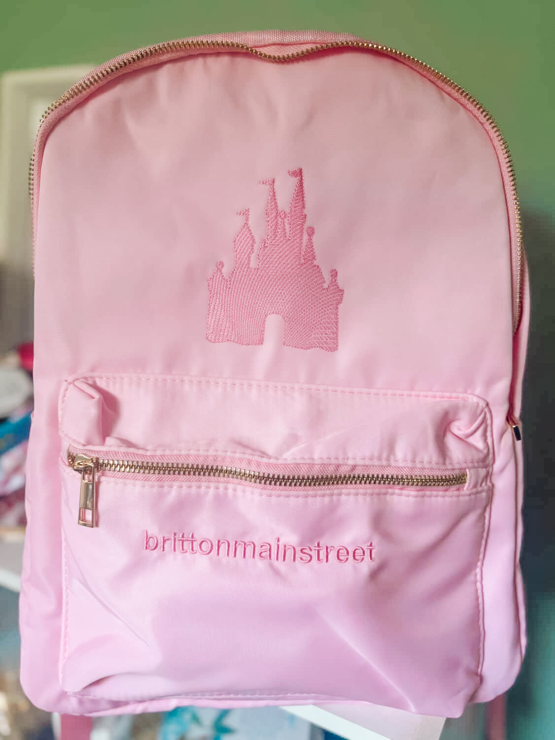 Pink backpack with castle design and 'brittonmainstreet' branding on a green background