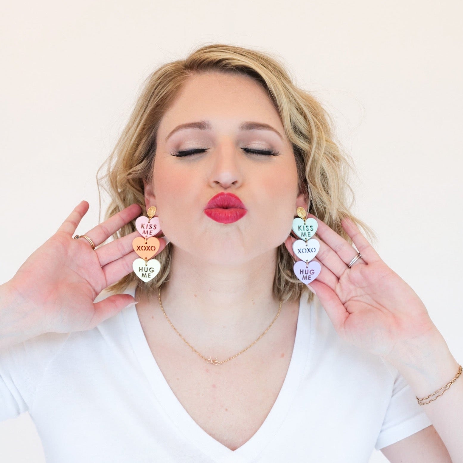 A woman wearing dangly three-tiered conversation heart earrings that read “kiss me,” “xoxo,” and “hug me,” on each earring from the top down.