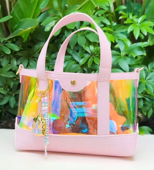 Pink handbag with transparent side panels showing colorful contents, against a green leafy background.