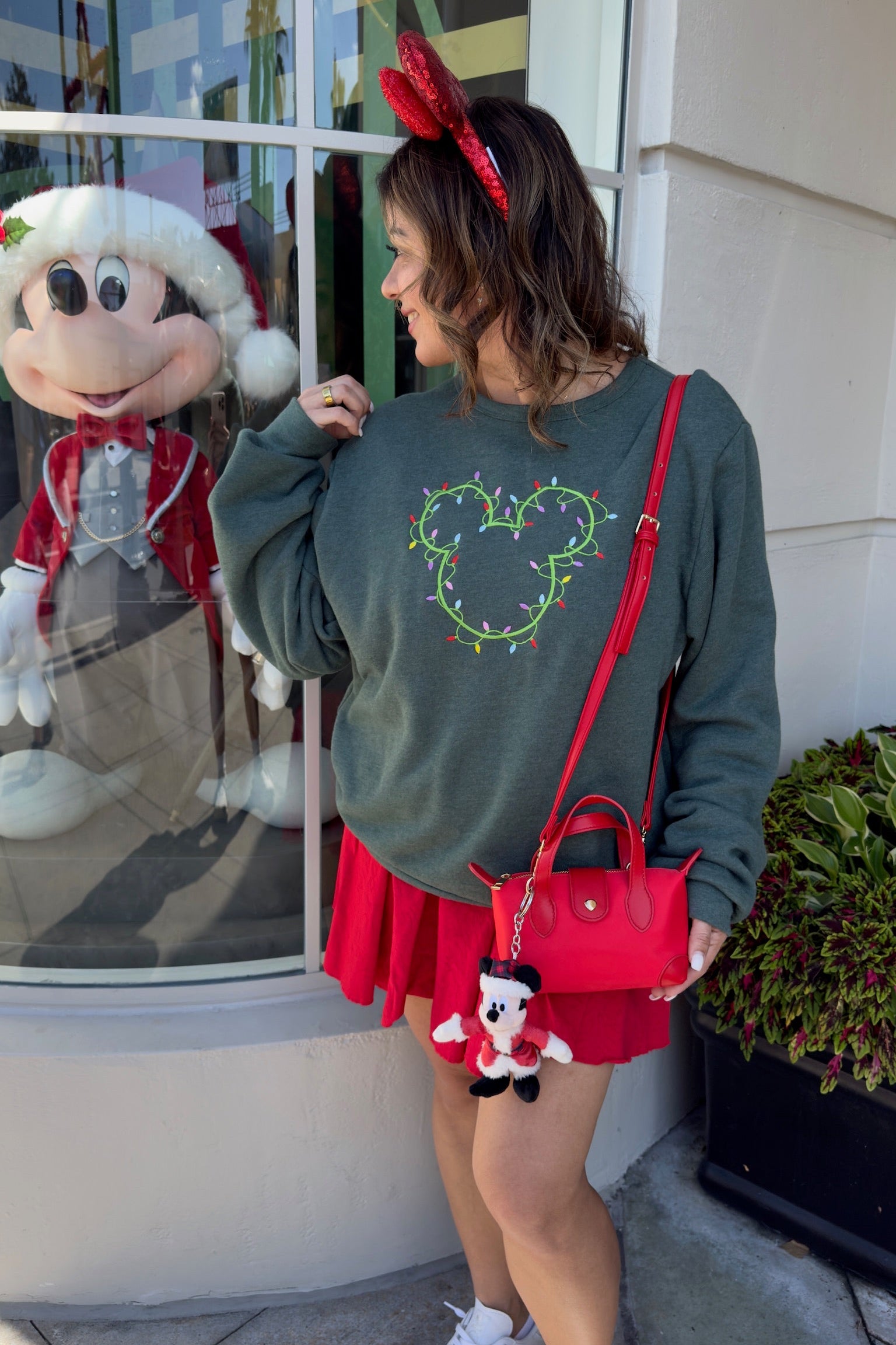 Woman wearing a festive outfit with a green sweatshirt and red skirt, standing in front of a store window with a character display.