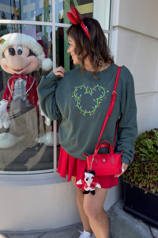 Woman wearing a festive outfit with a green sweatshirt and red skirt, standing in front of a store window with a character display.